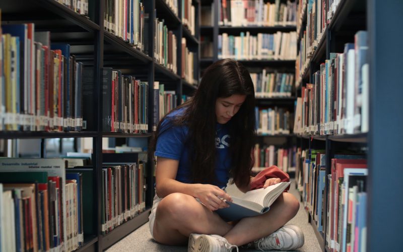 A young woman sitting on the floor of a library, reading a book intently.