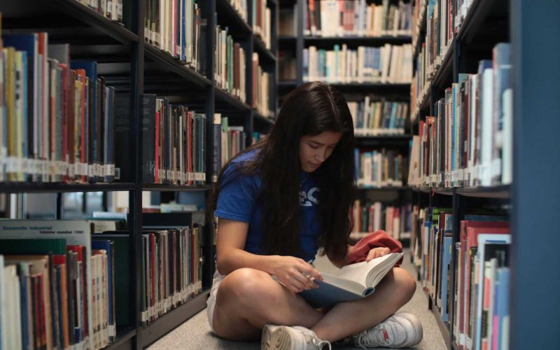 A young woman sitting on the floor of a library, reading a book intently.